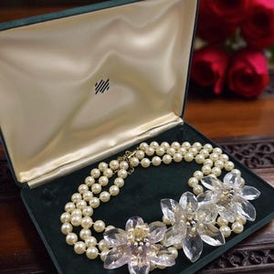 May include: A pearl necklace with three strands and clear flower accents rests in an open, green velvet jewelry box. The necklace is displayed on a dark wooden surface, with red roses in the background.