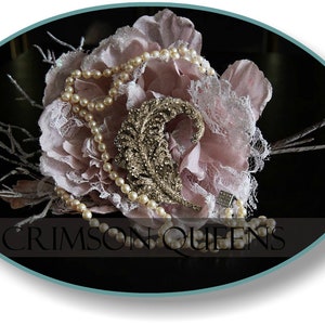 Op de afbeelding: Een roze en witte bloemstuk met een gouden veren broche en een parelketting. De opstelling is op een zwarte achtergrond met de tekst "CRIMSON QUEENS".