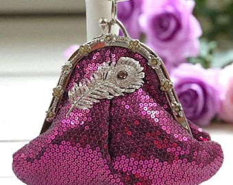 Può includere: Un piccolo portamonete con paillettes magenta con una cornice in metallo argentato. La borsa presenta una piuma decorativa argentata e una piccola gemma rotonda. La cornice è adornata da piccoli accenti floreali color oro.