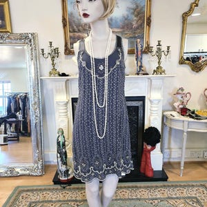 Peut inclure: Une robe de style flapper bleu marine avec des perles complexes et un ourlet festonné. La robe est présentée sur un mannequin, accessoirisée d'un long collier de perles et d'une pince à cheveux ornée de bijoux. La robe a un col rond et un design sans manches.