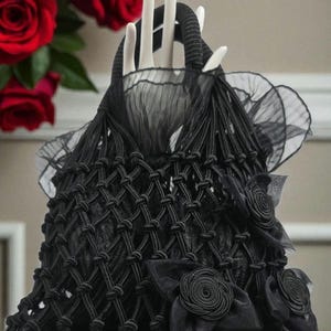 May include: Black macrame handbag with a knotted design, adorned with fabric rosettes and bows. The bag features a structured handle and a sheer, ruffled trim. The bag is displayed on a white surface.