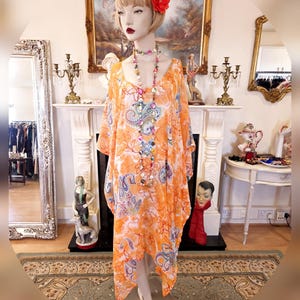 Peut inclure: Une longue robe à motif orange et blanc avec un col rond et une jupe asymétrique fluide. La robe est sur un mannequin avec une fleur rouge dans les cheveux et un long collier de perles.