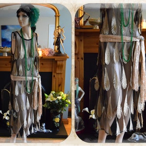 May include: A vintage-style dress on a mannequin, featuring a beige underdress overlaid with a sheer black fabric adorned with gold feather-like appliqués. The dress is accessorized with a beaded green necklace and a fringed belt. A matching headpiece completes the look.