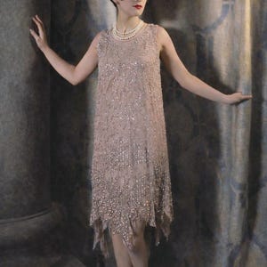 May include: A vintage-style, sleeveless, knee-length dress in a light tan colour, heavily embellished with sequins. The dress has a slightly asymmetrical hemline and is paired with matching heels. A pearl necklace completes the look.