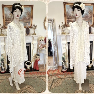 May include: A white lace dress with a beaded neckline and a matching white skirt. The dress is on a mannequin in a room with a fireplace and a mirror.