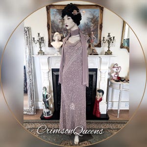 May include: A mauve, floor-length evening gown with detailed beadwork. The dress has a cowl neckline and a matching scarf. The mannequin is styled with a black wig and a floral hair accessory. The text "Crimson Queens" is at the bottom of the image.