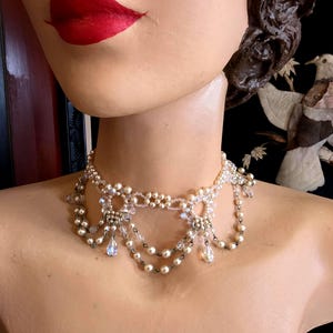 May include: A delicate pearl and crystal choker necklace with a layered design. The necklace features multiple strands of pearls and crystals, with a central focal point of a floral design. The necklace is a beautiful and elegant piece of jewellery.