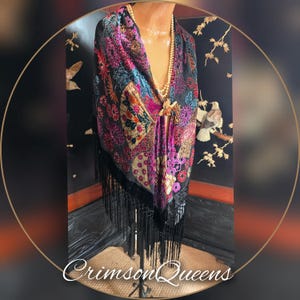 May include: A colorful shawl with a floral pattern in shades of pink, purple, and green, displayed on a mannequin. The shawl has a black fringe and is secured with a gold brooch. The text "Crimson Queens" is visible at the bottom.