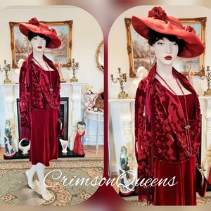 May include: A red velvet dress with a matching shawl and a red hat with a flower accent. The dress has a fitted bodice and a flowing skirt. The model is wearing a pearl necklace and a brooch. The image has the text "Crimson Queens" at the bottom.