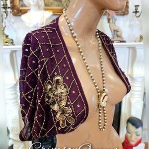 May include: A purple beaded shrug with gold sequins and a geometric pattern. The shrug has short sleeves and a V-neckline. The shrug is being worn by a mannequin with a pearl necklace.