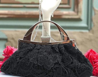 Può includere: Borsa a mano in pizzo nero con manico e cornice argentati. La borsa presenta un motivo floreale in pizzo e una tracolla a catena. Un manichino a mano bianco tiene una collana e un anello argentati, con rose rosse sullo sfondo.