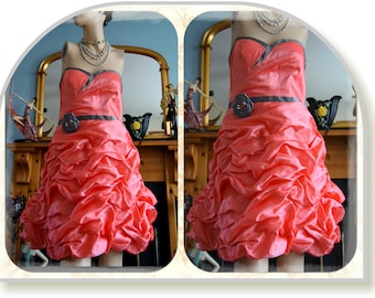 May include: A coral pink strapless dress with a black satin sash and a large black flower accent. The dress has a ruffled skirt and is made of a shiny fabric.