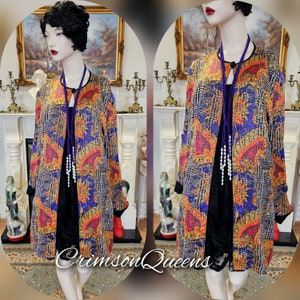 Könnte beinhalten: Eine langärmelige, gemusterte Jacke mit einem blauen, orangefarbenen und gelben Blumendesign. Die Jacke hat einen Stehkragen und einen Knopfverschluss. Das Model trägt ein schwarzes Kleid und eine violette Perlenkette.