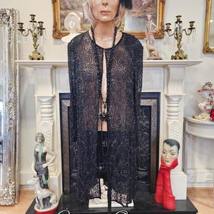 Peut inclure: Une robe de flapper noire à paillettes avec une superposition transparente et un collier de perles. La robe est sur un mannequin avec un bandeau noir et une plume. Le texte "Crimson Queens" est visible en bas de l'image.