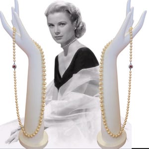 May include: A pearl necklace with a red gemstone clasp is displayed on a white mannequin hand. The necklace is composed of numerous small, round pearls. A black and white image of a woman wearing a pearl necklace is in the background.
