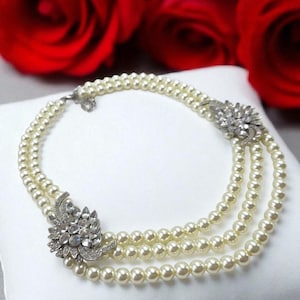 May include: A three-strand pearl necklace with decorative silver floral clasps. The necklace is presented on a white cushion, with red roses in the background. The pearls are a creamy off-white colour.