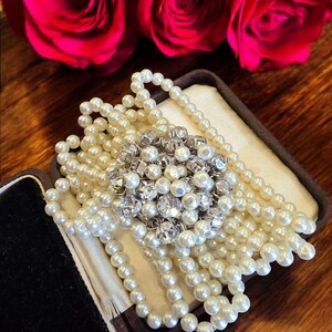 Può includere: Una collana di perle bianche con un grande spilla di strass scintillante in una scatola di gioielli in velluto marrone.