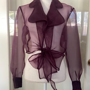 Peut inclure: Un chemisier en mousseline de couleur bordeaux avec un grand col et des manches longues. Le chemisier se noue à l'avant avec un nœud.