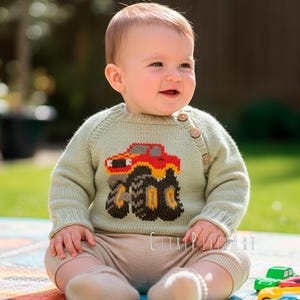 Puede incluir: Un bebé con un suéter verde claro con un diseño de camión monstruo rojo y amarillo. El bebé está sentado en una alfombra colorida con coches de juguete cerca. El suéter tiene tres botones de madera en el hombro.
