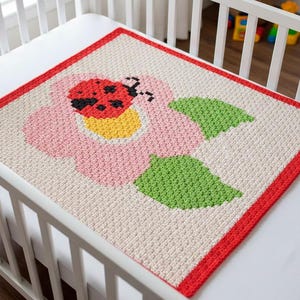 Puede incluir: Una manta de bebé hecha a mano con un diseño de mariquita y flor. La manta presenta una flor rosa con un centro amarillo y hojas verdes, un borde rojo y una mariquita. La manta está colocada sobre una cuna blanca.