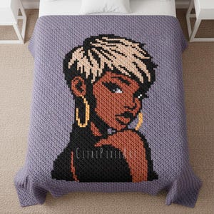 Puede incluir: Una manta lavanda muestra un retrato pixelado de una mujer con cabello corto con mechones rubios y aretes de aro dorados. La mujer tiene la piel morena y viste una blusa negra. La manta está sobre una cama con almohadas blancas.