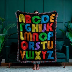 May include: A colorful alphabet blanket with each letter in a different color. The blanket is made of woven yarn and has a fringe border.