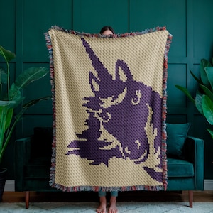 May include: A beige throw blanket with a large purple unicorn design. The blanket has a fringed edge with red, blue, and beige threads. The unicorn is depicted in a pixelated style, with a detailed horn and mane.
