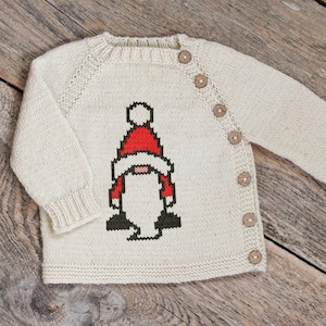 May include: Cream-colored knitted baby sweater with a button closure and a gnome design on the front. The gnome has a red hat and a white beard. The sweater is made of a soft, textured yarn.
