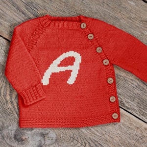 Peut inclure: Un cardigan rouge vif en tricot, orné d'un grand "A" blanc sur le devant. Il possède des manches longues, un col rond et des boutons en bois. Le vêtement est présenté sur une surface en bois.