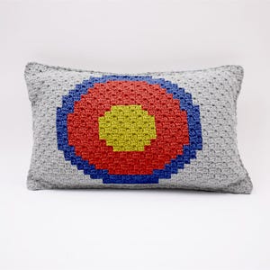 May include: A gray crocheted pillow with a colorful circular design. The circle is made up of blue, red, and yellow yarn.