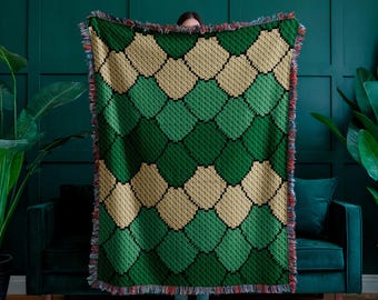 May include: A green and tan patterned throw blanket with a diamond design. The blanket has a fringed edge with red, blue, and white accents. The blanket is displayed in front of a dark green wall and a green sofa.