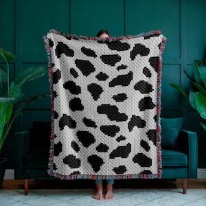 May include: A black and white crocheted throw blanket with a cow print pattern. The blanket has a fringe border.