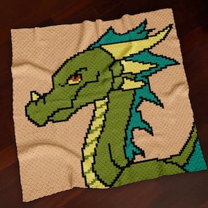 Op de afbeelding: Een handgemaakte gehaakte deken met een groen drakenontwerp. De draak heeft gele en blauwgroene accenten, met een beige achtergrond. De deken is gemaakt met een pixelachtig ontwerp, waardoor het een unieke, handgemaakte uitstraling krijgt.