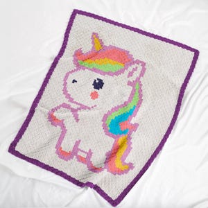 May include: A white crocheted blanket with a purple border featuring a unicorn with a rainbow mane and tail.