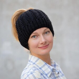 May include: A black knitted ponytail beanie hat. The hat is worn on the head, with the ponytail extending through the opening at the top. The beanie is made of a thick, textured yarn, and is designed to keep the head warm.