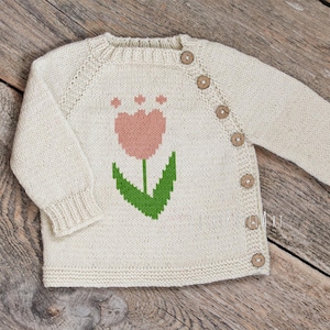 May include: Cream-colored knitted baby cardigan with a pink tulip design on the front. The sweater has long sleeves, wooden buttons, and ribbed cuffs. The garment is displayed on a rustic wooden surface.