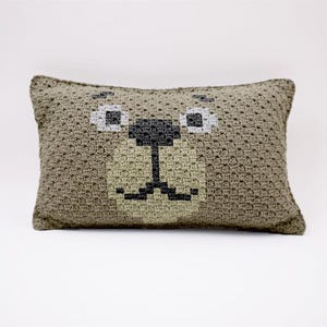 Puede incluir: Una almohada de crochet marrón con una cara de oso bordada en la parte delantera. El oso tiene ojos negros, una nariz negra y una boca marrón.