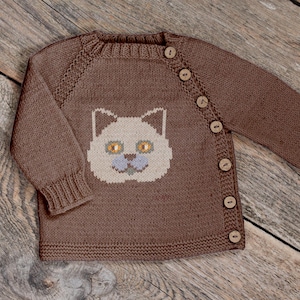 Puede incluir: Cárdigan de punto marrón con diseño de cara de gato. El cárdigan tiene una parte delantera abotonada y mangas largas. La cara del gato es beige con ojos amarillos y nariz y boca azules. La prenda está hecha de un material de punto suave.