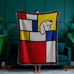 May include: A colorful throw blanket featuring a geometric design with a white dog illustration. The blanket has a red, yellow, blue, and white color scheme with a black border and fringe.