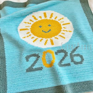Puede incluir: Una manta de bebé de ganchillo en tonos de azul, blanco, amarillo y gris. La manta presenta un diseño de sol sonriente en el centro y el número "2026" debajo. La manta tiene un borde gris.