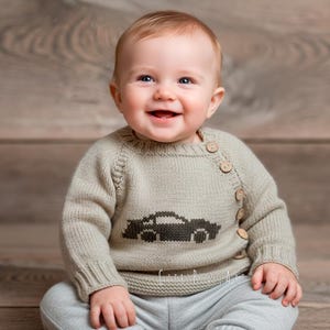 Könnte beinhalten: Ein lächelndes Baby trägt einen hellgrauen Pullover mit einem schwarzen Autodesign. Der Pullover hat Holzknöpfe an der Schulter und wird mit einer grauen Hose kombiniert. Der Hintergrund ist eine Holzoberfläche.
