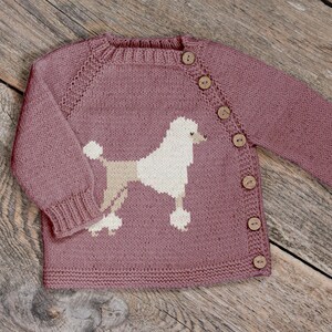 May include: A mauve knitted baby cardigan with a poodle design. The button-up sweater features a white and beige poodle graphic on the front. The long-sleeved sweater has wooden buttons and ribbed cuffs.