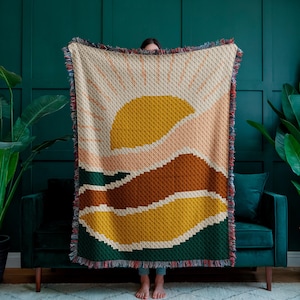 May include: A woven tapestry throw blanket with a colourful abstract design featuring a yellow sun, pink and brown hills, and green grass. The blanket has a fringe border.