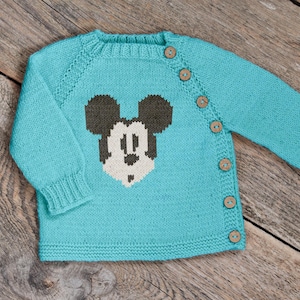 May include: A turquoise knitted baby cardigan with a Mickey Mouse design on the front. The cardigan has long sleeves, a round neckline, and wooden buttons. The Mickey Mouse design is in black, white, and beige.