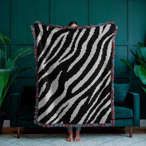 May include: A black and white zebra patterned woven throw blanket with fringe on the edges. The blanket is being held up in front of a person.