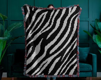 May include: A black and white zebra patterned woven throw blanket with fringe on the edges. The blanket is being held up in front of a person.