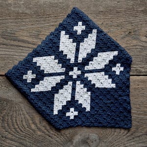 May include: A navy blue and white crocheted square with a snowflake design. The snowflake is composed of white stitches and features a symmetrical pattern with cross-shaped accents. The item is placed on a rustic wooden surface.