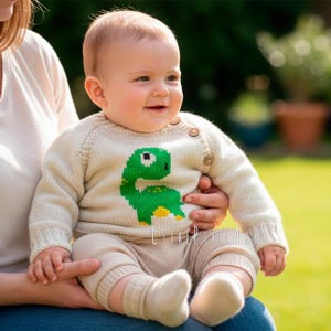 Może przedstawiać: Niemowlę w kremowym swetrze z dzianiny z zielonym wzorem dinozaura. Sweter ma drewniane guziki i jest połączony z pasującymi beżowymi spodniami i skarpetkami. Dziecko się uśmiecha.