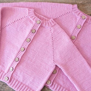 May include: Two light pink knitted cardigans with wooden buttons. The sweaters have long sleeves and ribbed cuffs and hems. The cardigans are made of a soft, textured yarn.
