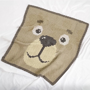 May include: A brown crocheted blanket featuring a bear face design. The bear has a brown nose, black eyes, and a beige mouth.